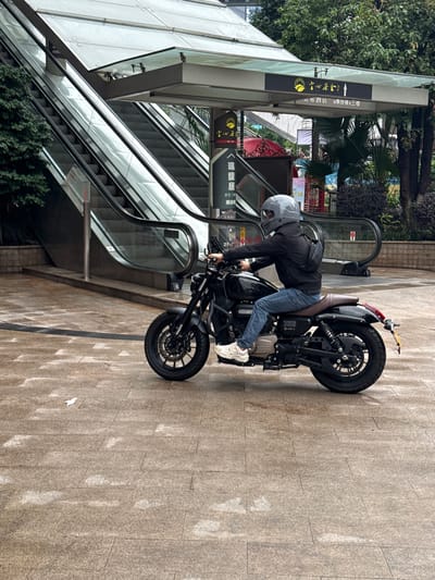 A motorcyclist in a grey helmet, dark jacket, jeans, and white sneakers is on a black motorcycle on a paved pedestrian walkway in Nan'an District, China, adjacent to an escalator leading to a building. A sign indicates directions to "珮馥居 茶艺" and "五市酒店 请乘扶梯三楼". The surrounding area includes trees and foliage. The photograph is oriented sideways.