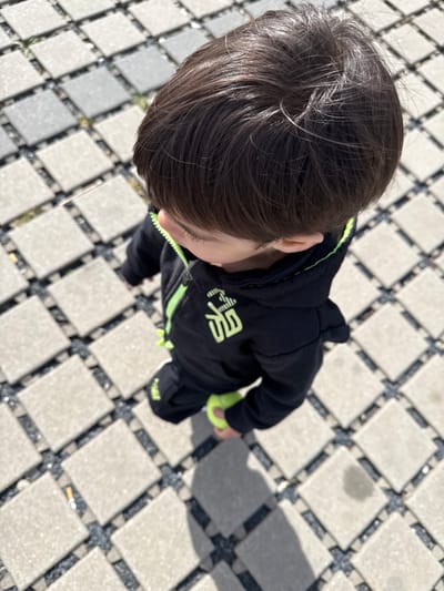 An overhead shot depicts a child standing on a paved surface. The child has dark hair and is wearing a black hooded jacket with green accents and a symbol on the chest. They are holding a lime green tennis ball in their right hand. The paving is made of square stones with dark filler between them. A cigarette butt is visible on the paving in the lower right of the image. The location is inferred to be Junglinster, Luxembourg, although there is no explicit information to confirm. The image is brightly lit, suggesting daylight conditions.