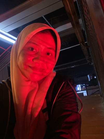 This media file depicts a close-up, low-angle shot of a person wearing a light-colored headscarf and a dark jacket. The scene is bathed in a strong red light, which casts dramatic shadows and highlights on the person's face. The person's eyes are open and looking towards the camera, with a slight smile on their lips, suggesting a relaxed or perhaps slightly playful mood. Their skin appears flushed due to the red lighting.
In the background, hints of an urban setting can be seen. There are blurred structures and lights, suggesting an outdoor environment, possibly at night or in a dimly lit area. The overall lighting and the presence of what appear to be building elements and outdoor lighting suggest an urban environment, potentially a street or an area with some form of overhead structure, like a walkway or a canopy. There are no discernible signs or text visible in the image. The time of day appears to be evening or night, given the dark background and artificial lighting. The weather is not discernible from the image.