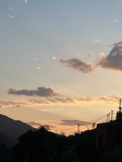The image depicts a view of Bin El Ouidane, Morocco, at dusk. The sky is filled with long streaks of clouds with reddish hues in the distance. There are electrical poles with connecting wires and a silhouette of a mountain ridge to the bottom-right. Some dark-colored, nondescript building can be seen to the right of the electrical poles. A dense stand of trees and vegetation occupies the lower right corner.