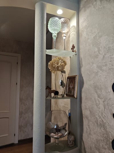 The image shows an interior scene. There is a decorative column on the left, with a wall behind it that has a textured, patterned finish. To the right of the column is a white shelf unit with several items displayed. The top shelf holds two clear glass hurricane candle holders with a textured pattern. Below that, on a glass shelf, is a clear vase with dried flowers, a small brown horse figurine, and a framed religious icon. The bottom shelf of the unit holds a large glass fishbowl filled with seashells. A small, light-colored figurine is placed next to the fishbowl. To the far left, a portion of a white door with vertical panels is visible. The overall impression is one of a home interior decorated with various ornamental objects. The image does not contain any information that would allow for determining its location within Barnaul, Russia.