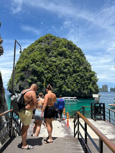 The image captures a scene at a pier in Ao Nang, Thailand, under a bright, partly cloudy sky, suggesting a warm, sunny day. The setting is a wooden pier with a railing, overlooking the turquoise waters of the sea. In the background, a lush, green, rocky island rises dramatically from the water, a common sight in the limestone karst landscape of the region. Several boats are visible docked at the pier or in the water, hinting at a tourist or transportation hub.
The foreground features several people, seemingly tourists, dressed in beachwear. One person in the front is wearing light yellow shorts and a backpack, facing away from the camera. Behind them, two individuals, a woman in a bikini and denim shorts and another woman in a bikini and black shorts, are also partially visible, facing the same direction. Further back, a person in a blue shirt with the word "SERVICE" clearly printed on the back is standing, possibly a staff member. The overall atmosphere appears relaxed and vacation-like, with people possibly waiting to board a boat or having just disembarked. The clear water and vibrant greenery of the island contribute to a tropical, scenic ambiance.