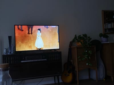 The scene depicts a dimly lit indoor living space in Lille, France. A large flat-screen television is centered on a dark wooden media console. The television screen displays an animated scene of a female character in a long yellow dress, standing on a light-colored wooden floor, with the lower extremities of other animated figures visible in the background. To the immediate left of the television, a white vertical PlayStation 5 console stands, accompanied by a smaller white electronic device. Below the television, a silver-colored stereo receiver with an array of buttons and a display is integrated into the media console. In the extreme bottom-left foreground, a clear glass jar with a cork stopper is partially visible. To the right of the media console, an acoustic guitar with a sunburst finish leans against a small, light-colored wooden cabinet featuring three drawers with white ceramic knobs. A potted plant with green and variegated leaves is situated on top of this cabinet. Behind the guitar and cabinet, a light-colored soft guitar case leans against the wall. Further to the right, a second potted plant in a purple pot rests on a higher surface. In the upper right portion of the frame, a framed mirror or shelf unit, possibly containing books, is mounted on the wall. The primary illumination in the room emanates from the television screen.