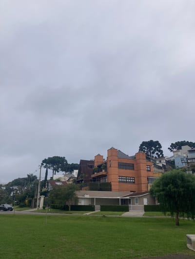 The image shows a view of a street in Curitiba, Brazil. It's a cloudy day. On the right is a green lawn adjacent to a sidewalk. Houses are visible along the street, with a mix of architectural styles, including a prominent brick building with multiple windows. A row of trees separates the houses from the lawn. In the lower portion of the frame, power lines and a parked car are visible. The general impression is of a quiet, upscale residential neighborhood.