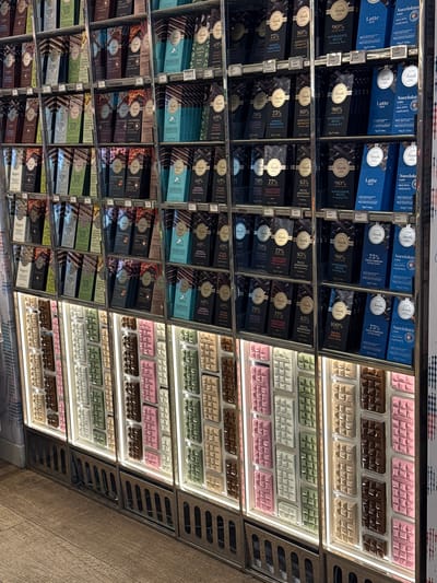 This photograph, taken indoors at an oblique angle, showcases a well-organized display of Venchi chocolate products in Noventa di Piave, Italy. The products are arranged on multiple shelves in a retail environment. On the left, the shelves are lined with packaged chocolate bars of varying percentages (60% to 100%) and flavors, including 'Latte', 'Nocciolata', and 'Bianco'. On the right, there are rows of unwrapped Venchi chocolate bars in pink, brown, white, and green hues. Price tags accompany each product, and the shelf structure is metal, complete with cooling vents. The lighting is bright and artificial. The setting is a typical retail space for chocolates.