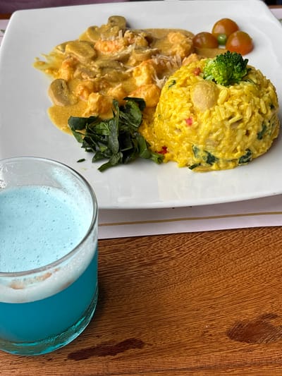 The image shows a plate of food on a white surface, next to a glass of blue liquid. The food consists of a creamy shrimp and mushroom dish, a mound of yellow rice with greens and broccoli, and a few cherry tomatoes. The blue liquid in the glass appears to be a beverage, possibly a smoothie or cocktail. The setting is likely a restaurant, given the presentation of the food and drink. The image is taken from a slightly elevated angle, looking down at the plate and glass. The background is blurred, but suggests a wooden table.