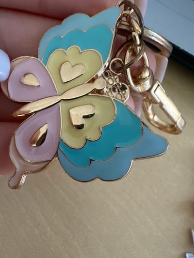 A close-up shot features a gold-tone keychain with a multi-colored butterfly charm. The charm is decorated with enamel in pastel shades of pink, yellow, and turquoise, with gold accents forming hearts and wing details. The butterfly is attached to a gold chain with a clasp. The background is a blurred wooden surface. No specific location in Murcia, Spain, is identifiable.
