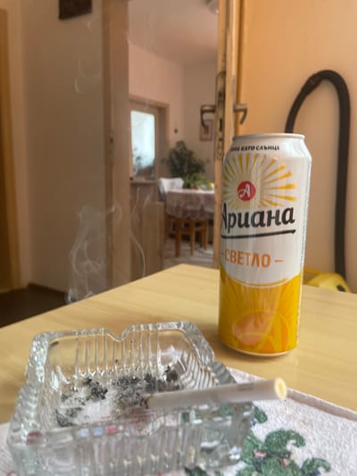 The image shows a can of Ariana Svetlo beer and an ashtray with a lit cigarette on a table. Smoke is visible in the air. The setting appears to be indoors, with a doorway leading to another room visible in the background. The location is not discernible as Elena, Bulgaria based on the provided image.