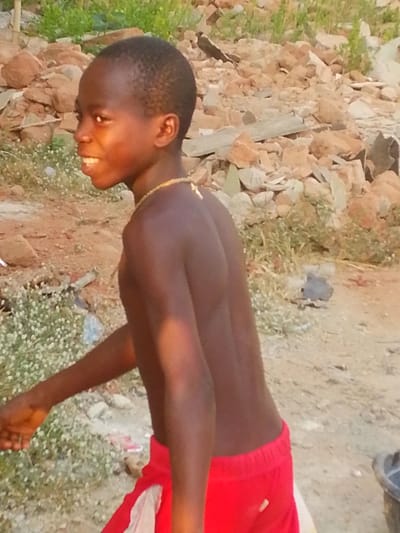 A young male child, estimated to be between 5 and 10 years old, with dark skin and short, dark hair, is depicted in a brightly lit outdoor scene in Enugwu-Ukwu, Nigeria. He is shirtless, wearing red shorts with a visible white panel on his left side, and a light-colored, possibly beaded, necklace with a small pendant. The child is captured in a dynamic posture, facing towards the viewer's left with his body slightly turned, his left arm extended forward. He exhibits a wide smile, revealing teeth. The immediate ground is reddish-brown earth with patches of small, white-flowered ground cover. The background consists of uneven terrain covered with various sizes of broken rocks, concrete debris, and sparse green vegetation. A dark-colored bucket or container is partially visible in the lower right corner. The lighting suggests natural daylight.