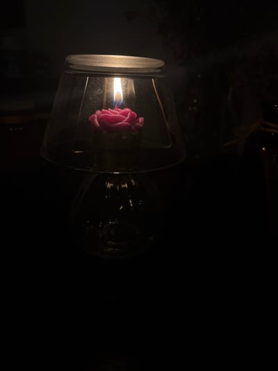A close-up shot of a rose-shaped candle burning within a clear glass bell jar, placed on a darker, possibly glass, base, in a dimly lit environment. The flame of the candle is bright and casts a warm glow on the surrounding petals of the candle. The glass bell jar has a slightly dusty or smudged appearance. The background is dark and out of focus, with hints of other objects or textures suggesting an indoor setting, possibly a home or a shop. The overall mood is serene and intimate. This image was taken in Ipswich, United Kingdom.