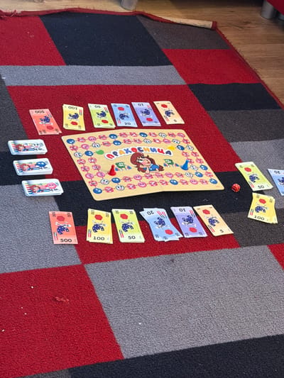 The image shows a child's board game, seemingly "Max Mütze", laid out on a red, grey, and black patterned rug. The board game is surrounded by play money of various denominations, including 5, 10, 20, 50, 100, and 500. A red die is placed near the game board. Stacks of small playing cards are positioned at the bottom of the image. The rug sits on a wooden floor. The location is not discernible from the image.