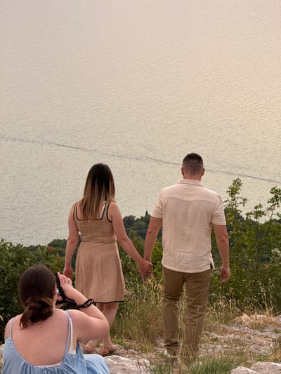 In Kamenovo, Montenegro, two individuals, a woman and a man, are pictured from behind, holding hands as they walk away from the viewer. The woman wears a sleeveless, light-colored dress, and the man is dressed in a light-colored short-sleeved shirt and brown pants. To the left, another person, also seen from behind, is seated and holding a camera, appearing to photograph the couple. They are wearing a blue top and white bra. The group is positioned on a rocky, grassy incline overlooking a body of water, likely the Adriatic Sea. The water is calm with visible ripples, and the horizon is blurred due to distance and atmospheric conditions, suggesting it is either early morning or late afternoon. Lush green foliage and trees surround the rocky terrain.