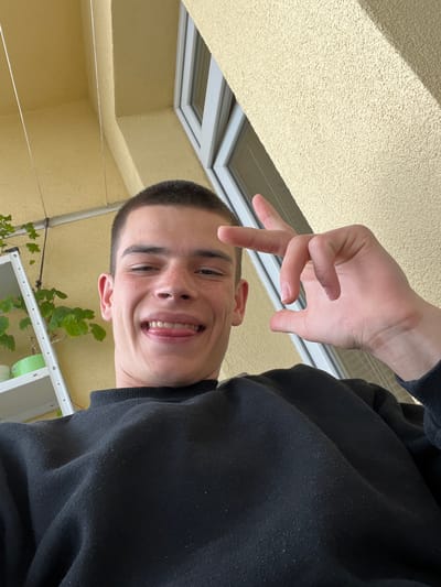 A close-up, downward-angled selfie depicts a young man with a buzz cut hairstyle, fair skin, and freckles. He is smiling and making a "peace" sign with his right hand. He wears a black long-sleeved garment. The background shows a yellow textured wall with a window frame visible at the top left. Below the man's head, there is a white structure with green foliage. The scene appears to be outdoors, potentially in a courtyard or balcony setting. There is no indication of the city of Sopot, Bulgaria.