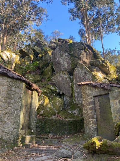 The image shows a rock formation with moss and vegetation growth in Esposende, Portugal. A man-made stone structure, possibly a stairway or wall, is integrated into the rock face. Trees are visible in the background against a blue sky. The light suggests it is daytime. No people are visible.