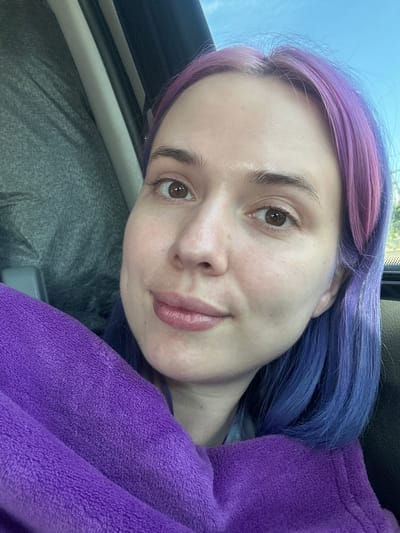 A selfie depicts a light-skinned person with dyed purple hair, the roots being more pinkish and the ends more blue, draped in a purple, plush blanket. The person gazes into the camera. The backdrop reveals the interior of a car, including portions of the window, door frame, and grey fabric. Based on the sky visible through the car window, it is daytime. The location of Pourrières, France, is not discernible from the visual elements of this image.