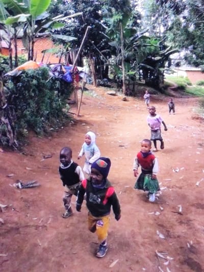 Seven children, ranging from toddler to elementary school age, are depicted moving along an unpaved, reddish-brown dirt path in a semi-rural area of Dodoma, Tanzania. The children are dressed in casual attire, including sweaters, shirts, skirts, and pants; one child in the immediate foreground wears a multi-colored knit cap. Three children are prominent in the foreground, advancing towards the viewer with smiles or engaged expressions. Two additional children are in the mid-ground, and two more are further down the path in the background. The path is bordered by dense green vegetation, including banana trees and various bushes. To the left background, a brick-colored building is partially visible, and another structure appears distantly on the far right. A makeshift structure, constructed from poles and draped with fabric, stands amidst the foliage on the left side of the path. The scene is illuminated by soft, diffused light.