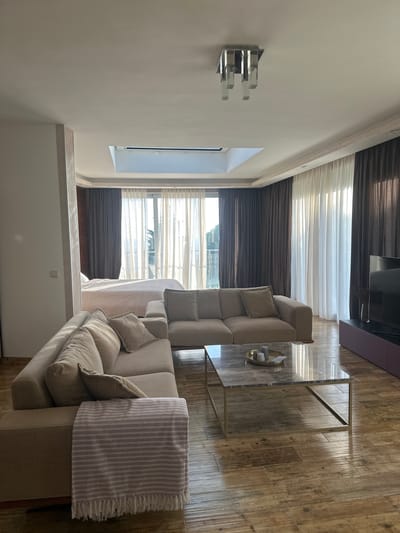 The image depicts a well-lit, modern living area and bedroom combined in what appears to be a hotel suite or apartment in Brca, Montenegro. In the foreground, a large, beige sectional sofa with multiple throw pillows is arranged in an L-shape. A light-colored throw blanket with pink and white stripes is draped over the arm of the sofa. In front of the sofa is a rectangular coffee table with a marble top and a gold metal frame, holding a decorative tray with candles.
The room opens up to a bedroom area with a bed covered in a light pink comforter. Behind the bed, large windows are covered by a combination of sheer white and dark brown curtains, allowing natural light to stream in. A skylight is visible in the ceiling above the windows, further illuminating the space. To the right of the living area, a large flat-screen television is mounted on a dark purple stand. The flooring throughout the visible area is made of wooden planks, reflecting the light. A contemporary ceiling light fixture with rectangular glass elements hangs from the ceiling. The overall impression is one of comfortable, stylish living with a view of the outdoors, characteristic of accommodations in coastal Montenegrin towns like Brca.