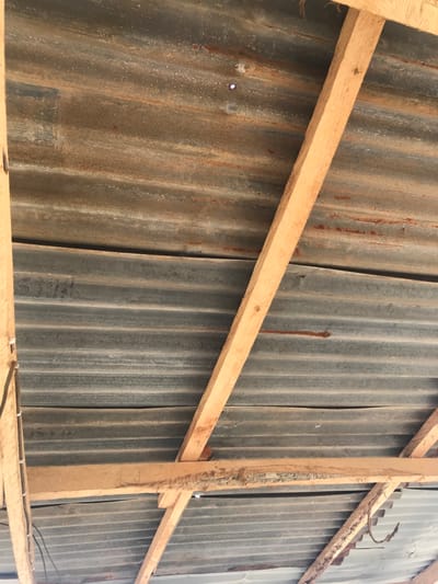 The image displays the underside of a roof structure situated in Oba-Ile, Nigeria. No individuals are present. The primary roofing material consists of corrugated metal sheets, exhibiting a mottled grey and oxidized reddish-brown coloration from extensive rust and weathering. These sheets are supported by an exposed framework of light-brown wooden beams, some displaying rough-cut surfaces and splintering. A distinct circular aperture in an upper corrugated sheet allows a visible point of external light to penetrate. Black insulated electrical wiring is secured vertically along a wooden beam on the left side of the frame. A single bent, rusted metallic object, consistent with a hook or fastener, is partially visible in the lower right quadrant.