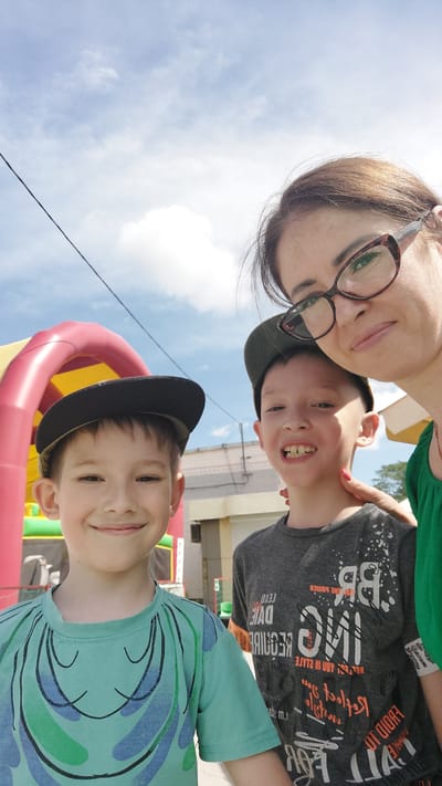 This is a selfie taken outdoors with a bright blue sky and scattered clouds. A woman wearing glasses and a green shirt is on the right. To her left are two young boys, both wearing baseball caps. The boy in the center has a gray t-shirt with white text. The boy on the left is wearing a teal t-shirt with a smiling face design. Behind them, there is a glimpse of a red and yellow inflatable structure, possibly a bouncy castle. The scene appears to be a daytime outdoor gathering or event. This image was captured in the city of Komsomolsk-on-Amur, Russia.