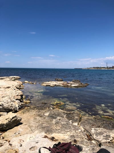 A vertical, eye-level, medium-wide shot captures a rocky shoreline on a bright, sunny day. The foreground is dominated by textured, light-colored rocks, some large and others smaller, with patches of brown seaweed. A dark red backpack and a black sandal rest on the rocks in the lower right corner. The middle ground features a rocky outcrop in clear, dark blue water, with a few small figures visible on the rocks. The background consists of the vast, dark blue sea meeting a clear, pale blue sky with a few wispy clouds. In the distance, along the horizon, is a faint line of buildings and industrial structures, suggesting the presence of a town or port. The image is located in the vicinity of Ароматненское сельское поселение, Ukraine.