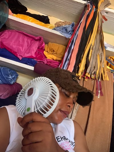 In Yelwa, Nigeria, a woman with dark skin wears a brown baseball cap and a white t-shirt. She is holding a white portable fan close to her face, her eyes are closed. She appears to be resting in a room filled with rolls of fabric and a bundle of colorful zippers hanging from the ceiling. The shelves are stacked with various textiles, including pink, blue, yellow, and maroon velvet materials, as well as a piece of sequined fabric. The environment suggests a workspace or storage area for a tailor or fabric vendor.
