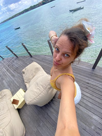 A woman is taking a selfie on a wooden pier in Nusa Penida, Indonesia. She has brown hair with a white flower tucked behind her ear, blue eyes, and is wearing a yellow bikini top and white bottoms. She is leaning on her hand, with her arm bent at the elbow, and her legs are extended. In the background, there is a clear blue ocean with several small boats and a distant coastline with buildings. The pier has a rope railing and two large beige beanbag chairs, one of which she is sitting next to. A small, light-colored wooden table with a dark wooden plaque is situated between the beanbag chairs.
