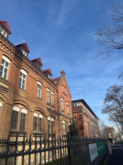 This daytime image, viewed at an upward angle, features a distinctive brick building under a clear, bright blue sky with faint wispy clouds. The main building, constructed of red-brown bricks, exhibits intricate architectural details, including decorative patterns in the brickwork. It has multiple floors with numerous windows, some arched and some rectangular, all framed in white. The roof, which appears dark red or brown, is adorned with several dormer windows, and a prominent ornate gable rises towards the top.

To the right of the foreground building, a black, decorative metal fence runs parallel, separating the building from a path or street. Beyond this fence, a taller, plainer brick building is partially visible in the background. Between the fence and the background building, a green and white sign is clearly visible, bearing the German text: "Schülerhilfe" at the top, and "Nachhilfe + Beratung" below it, indicating a student tutoring and advice service. Bare tree branches extend into the sky from the upper left corner, and more bare trees are visible in the background. The bright sunshine casts distinct shadows, highlighting the textures and architectural features. The scene suggests an urban setting on a clear day, likely in late autumn or winter due to the bare trees.