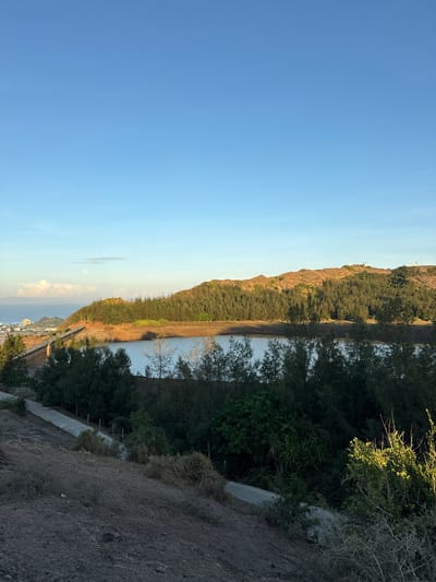 The image captures a scenic view of a reservoir on Lý Sơn Special Administrative Region, Vietnam, under a clear blue sky. In the foreground, a dusty, dry slope leads down towards a paved path winding through sparse vegetation. Beyond the path, a dense line of dark green trees forms a natural barrier around the reservoir. The reservoir itself is a calm body of water, reflecting the light of the sky and appearing somewhat murky. A dam structure, partially visible on the left, stretches across the scene, connecting to a distant coastline. The background features rolling hills covered in a mixture of trees and dry, reddish-brown earth. On the far left, a town with several buildings is nestled near the sea, which stretches to the horizon. A faint moon and a few wispy clouds are visible in the upper left quadrant of the sky. The overall impression is one of a serene natural landscape with hints of human infrastructure.