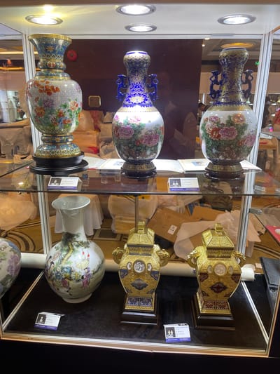 The image displays a collection of decorative porcelain vases and golden ornate objects, likely exhibited in a retail or museum setting. The items are displayed vertically within glass cases, with spotlights illuminating them from above.
The subjects include several large porcelain vases. One prominently featured vase is predominantly blue with intricate gold patterns and what appear to be dragon motifs. It is adorned with colorful floral designs, featuring pink and white peonies. Another vase, positioned below the blue one, shares a similar shape and is decorated with a detailed floral pattern that includes pink peonies, green leaves, and hints of orange and red. Further down, a third vase showcases a delicate painting of figures in a landscape. The golden objects are ornate, box-like structures with intricate carvings and decorative inlays, suggesting a decorative or perhaps functional purpose, like containers or pedestals.
The scene appears to be indoors, within a brightly lit display area. The background shows reflections in the glass, suggesting other display cases or the general environment of an exhibition hall. There are also glimpses of cardboard boxes and other materials in the background, indicating a potential retail or exhibition setup. The overall impression is one of refined craftsmanship and artistic display.
Visible text includes labels on the display items and potentially on the boxes. One label, positioned on a white card, has Chinese characters and an image of a vase. Another purple label also contains text in what appears to be Chinese.
The lighting suggests an indoor environment, possibly during the day or with artificial lighting. There are no clear indicators of weather or a specific time of day, beyond the artificial lighting. The overall mood is one of appreciation for fine art and craftsmanship. The location context of Futian District, China, aligns with the style and nature of the displayed items, which often reflect traditional Chinese artistry.