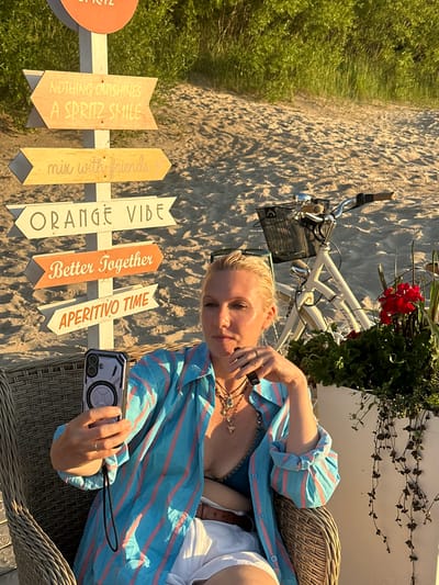 A woman is seated in a wicker chair on a sandy beach, holding a phone. She is wearing a blue and pink striped shirt and a blue bikini top, with a necklace around her neck. She has blonde hair and is looking down at her phone. Behind her, a bicycle is leaning against a planter filled with red flowers and greenery. A signpost with several directional arrows is visible to her left. The arrows have text that reads: "NOTHING OUTSHINES A SPRITZ SMILE", "mix with Friends", "ORANGE VIBE", "Better Together", and "APERITIVO TIME". The sun is shining, casting shadows on the sand, suggesting it is daytime. The scene appears to be a relaxed, summery setting.