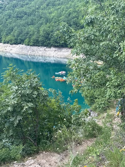A high-angle, medium shot captures a vibrant turquoise lake bordered by a densely forested hillside in Rudinice, Montenegro. The water is calm, reflecting the lush green trees that dominate the background and foreground. Several colorful kayaks and paddle boats are scattered across the lake, some with people paddling. The shoreline is a mix of rocky terrain and dense vegetation. The overall scene conveys a sense of natural beauty and recreational activity.