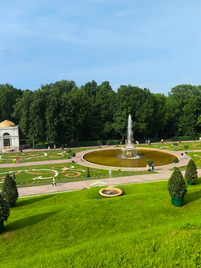 The image depicts a view of the Peterhof Palace and Gardens in Peterhof, Russia. In the foreground, a lush green lawn slopes downwards. In the middle ground, a circular pond with a large fountain is situated, surrounded by pathways and manicured gardens featuring geometric patterns of grass and colored material. To the left of the pond is a white pavilion with a golden dome. Further back, a dense line of mature green trees forms a backdrop. Several people are seen walking along the pathways in the gardens. A smaller, simpler fountain is visible on the lawn to the left of the main pond. The sky above is clear and blue.