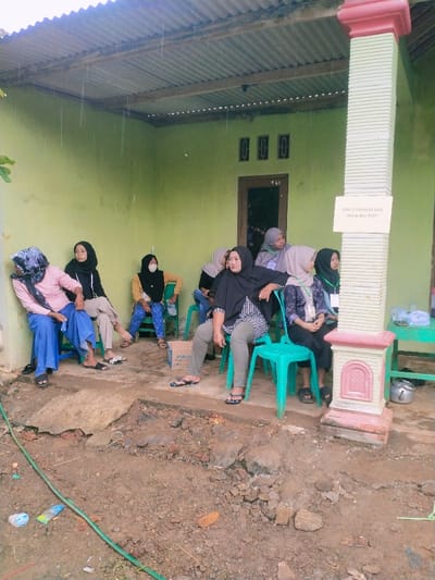 The image shows a group of seven women sitting on chairs under a covered porch in Kecamatan Kedawung, Indonesia (latitude: -7.493, longitude: 111.07). They are all wearing headscarves and traditional clothing. Some women are wearing face masks. One woman is sitting on a blue plastic chair and is wearing a pink shirt and blue pants. Another woman is sitting on a green chair and is wearing a black shirt and brown pants. The woman on the left is holding a handbag. The porch has a green painted wall with a window and three small windows above it. The porch is covered with a corrugated metal roof. The women are sitting in front of a pillar that has a sign that reads "JAMA' S TOHUN KE ATAS BOLEH BILA TIKET." The ground is dirt and there is a green hose running across the ground in the foreground. The scene appears to be informal and casual.