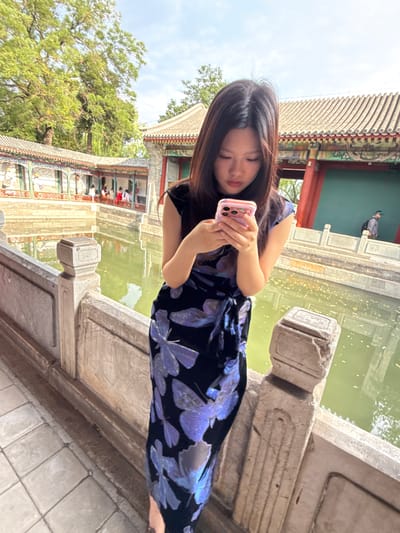 This media file depicts a young woman holding a pink phone, engrossed in its screen. She has long, dark hair and is wearing a dark blue dress adorned with a pattern of purple butterflies. The woman is standing on a stone bridge or walkway, with her body angled towards the camera.
The background reveals a traditional Chinese architectural setting. To the left, there's a building with a curved, tiled roof and ornate decorative elements. Green trees are visible behind the building. To the right, a body of water, likely a pond or lake, reflects the greenish hue of the surroundings. A person in a dark jacket is visible on the edge of the water in the distance. The overall scene suggests a serene, possibly historic, outdoor location. The lighting indicates it might be daytime, with a cloudy sky.
The scene appears to be in Xicheng District, China, given the architectural style and the context provided. The woman's focus on her phone suggests she is engaged in a personal activity, possibly taking a photo or browsing. The setting evokes a sense of tranquility and cultural richness. No visible text is present in the media.