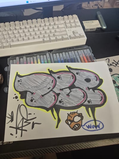 The image shows a white keyboard on a black desk. To the right of the keyboard is a pack of HEETS cigarettes and a silver vaping device. In front of the keyboard, a white piece of paper with graffiti art is laid out. The graffiti features large, puffy gray letters outlined in black and pink, with a yellow glow around them. Below the graffiti, there is a signature in black ink and a small drawing of a cartoonish face with its mouth open, along with a speech bubble containing the word "Wow" in blue. A set of markers is visible behind the paper. The background shows a portion of a computer monitor. The scene appears to be in a home or office setting. There is no indication of the city of Novi han, Bulgaria in the image.