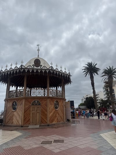 The image captures the Quiosco de la Música, a wooden bandstand located in Arrecife, Spain. The structure features a distinctive dome, decorative elements, and is positioned on a patterned red and grey brick paved area. Two tall palm trees rise in the background, along with a building. Adjacent to the bandstand, a crowd of people stand and a stage with sound equipment is visible. The sky is overcast with grey clouds.
