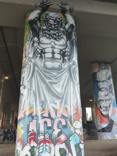 The image displays two cylindrical concrete pillars, likely part of an overpass or bridge structure, covered in vibrant graffiti. The pillar in the foreground features a large, monochromatic mural of a muscular, bearded male figure in classical drapery, rendered in shades of grey and white. The figure appears to be in distress, with arms raised and hands outstretched. Below this figure, the pillar is adorned with colorful graffiti lettering and cartoonish figures, including a shark with sharp teeth and a stylized face with wide eyes. The second pillar, to the right and slightly further back, is also covered in graffiti. It prominently features a black and white illustration of a face with sharp angles and a purple mark on its cheek, against a background of blue and orange hues. The ground surrounding the pillars is sandy or dirt, with some scattered debris. In the background, a metal fence with red vertical posts is visible, along with more graffiti on a lower wall. A street lamp with an illuminated bulb is positioned on the left side of the frame. The scene appears to be in an urban outdoor setting, under the shade of a concrete structure. While the description of the graffiti is detailed, there is no specific visual information that allows for the identification of the city of Kaliningrad, Russia.