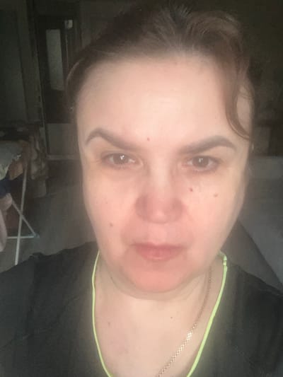 This is a close-up, head-and-shoulders selfie of a woman, taken from a slightly high angle and rotated so that her head is tilted. She has light skin with a few small red blemishes on her cheeks and nose. Her eyebrows are dark and well-defined. Her eyes are open, and she appears to be looking directly at the camera with a neutral or slightly somber expression. Her lips are slightly parted and appear to have a subtle pinkish tint. Her brown hair is pulled back, with some strands falling forward. She is wearing a black top with a neon yellow trim around the neckline, and a delicate gold chain necklace is visible. The background is dark and out of focus, with some indistinct shapes that suggest an indoor setting, possibly a room with shelves or furniture. The lighting is somewhat dim, creating a soft, diffused effect. There is no visible text in the image. The context provided indicates the location is Tula, Russia.