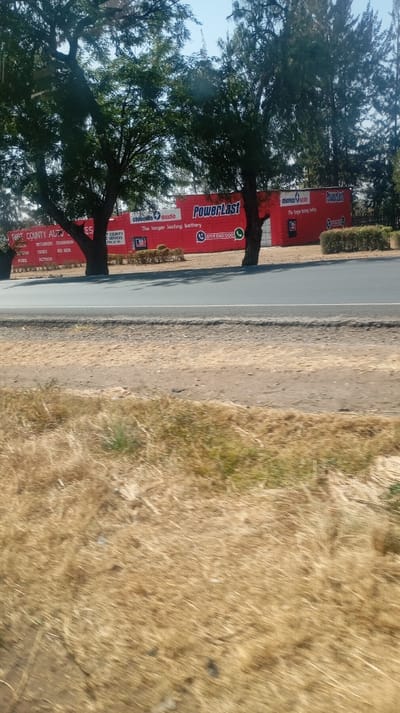 A roadside view features a large, vibrant red banner or wall advertising "PowerLast" batteries from "chloride exide."  The slogan, "The longer lasting battery," is prominently displayed. A phone number, "0719 030 000," and a WhatsApp icon are included.  To the left, partially obscured by trees, is a sign for "First County Auto," listing brands such as "Mitsubishi," "Isuzu," and "Ford." A gray road separates the advertising from a field of dry, golden grass. The sky is a pale blue.