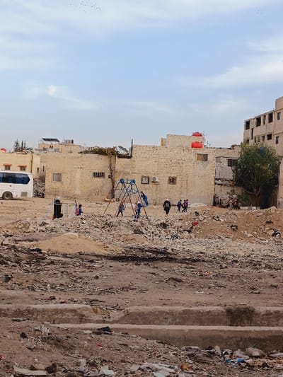 The image depicts an outdoor scene in the Kafr Sousa Municipality, Syria, characterized by a desolate, unpaved urban lot. The foreground is composed of uneven, reddish-brown earth mixed with construction debris, rubble, and scattered refuse, including sections of concrete. In the midground, a rudimentary playground features a blue metal climbing frame or swing set. Several individuals, primarily children, are present near this structure. Specifically, approximately two children are near the blue playground, with one appearing to interact with it. A figure in dark clothing walks towards the right, away from the playground, followed by a small group of children. To the far left, near a black stand-like object, two indistinct figures are visible. Piles of light-colored earth or sand are also scattered across this area. The background features a collection of buildings constructed from light-colored masonry blocks, some appearing complete with windows, while others are partially built or in disrepair. Red water storage tanks are mounted on the rooftops of several completed structures. A white bus or large van is parked on the left side of the background. Sparse green vegetation, including small trees, is visible among the buildings. The sky is light blue with faint, diffuse cloud cover, and a flock of small birds is observed in the upper frame. The lighting suggests daytime conditions.