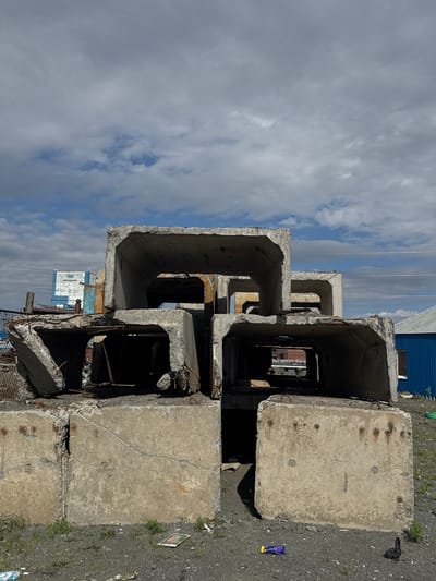 The image depicts a stack of large, weathered concrete pipes, likely in an industrial or construction area. The pipes are arranged in a staggered formation, creating a visually interesting pattern. The top of the pipes and the surrounding ground are visible. The ground appears to be covered in gravel and some sparse vegetation. In the background, there is a blue corrugated metal building and a white building with some blue accents. Power lines are also visible against a cloudy sky. The lighting suggests it is daytime, and the clouds indicate potentially overcast or partly cloudy weather. There are no people visible in the image. One of the pipes has some debris inside and exposed rebar, suggesting it may be damaged or undergoing demolition. A small purple and yellow wrapper is visible on the ground to the right of the pipes, and a black plastic bag is also present. No legible text is visible in the image. The scene suggests an outdoor, possibly remote or industrial, location in Norilsk, Russia.