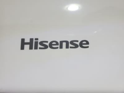 The image is a close-up, eye-level shot of the word "Hisense" in black, bold, sans-serif font, centered on a plain white surface. The surface appears to be a product casing, possibly of an appliance. There is a bright, circular light source reflected in the upper portion of the white surface, casting a slight glare. The overall lighting is even, with no shadows. The background is entirely white and featureless, except for the light reflection. The image does not provide any information about its location in the city of Unknown, Nigeria, or details about people, actions, or interactions.