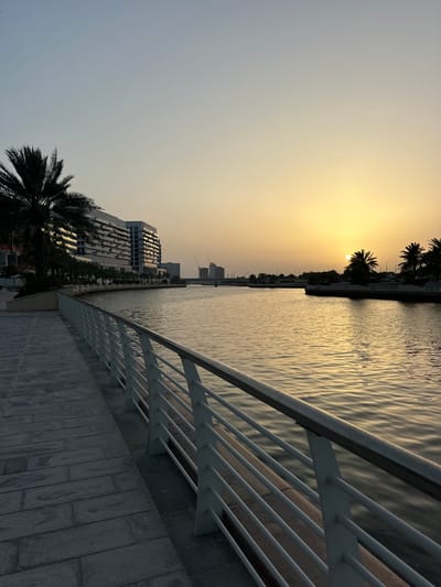 The image depicts a waterfront scene in Abu Dhabi, United Arab Emirates, during a sunset or sunrise. The sun is low on the horizon, casting a golden light over the water. A white railing runs along the edge of a paved walkway. Palm trees are visible along the shoreline. In the distance, modern buildings and construction cranes are visible. The water shows ripples and reflections of the sunlight. The sky transitions from a light blue or grey to a yellow or orange hue near the horizon.