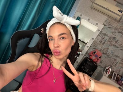 The image shows a young woman with long, dark hair and a white fluffy headband. She is wearing a bright pink top and is holding up two fingers in a peace sign. She is leaning into the camera and making a pouty face with her lips. In the background, there is a mirror with lights, an air conditioner, and some stuffed toys. The room appears to be a bedroom or a dressing area. The woman is sitting in a black gaming chair, and there are teal-colored curtains in the background. The image seems to be a selfie taken indoors, likely in the evening or at night, as the lighting is artificial. The woman's expression is playful and relaxed. There is no visible text in the image, except for a small logo on the air conditioner which appears to be "SAMSUNG".