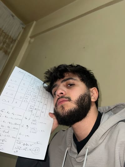 A young man with dark hair and a beard holds a piece of paper with a table and handwritten calculations. The table has columns labeled "المضاع" (multiples) and A, B, C, and D. Rows are labeled A-B, D-C, C-D, and D.A. The table contains numerical data. Below the table, there are equations including variables like RA, RC, RD, and VA, as well as calculations involving Σ and V. A note appears to the right, mentioning ΔH.