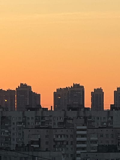 This is a photograph of the skyline of Sinyavino, Russia, during sunset. The sky is a gradient of orange and yellow. Silhouetted against the sky are several apartment buildings, some appearing taller and more modern than others. The foreground shows a closer view of more densely packed, older apartment buildings with visible balconies and windows. The overall impression is one of an urban residential area at dusk.