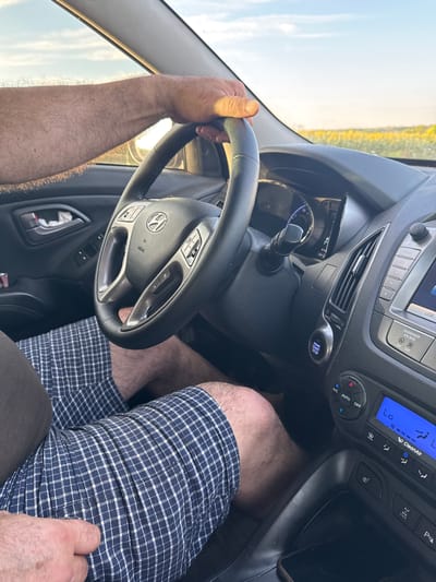 The image shows the interior of a car, with a person's arms and legs visible. The person is wearing checkered shorts and has their hand on the steering wheel. The car is likely in motion, as indicated by the blurred background which shows a field of yellow flowers under a bright sky. The dashboard of the car is visible, including the steering wheel, instrument cluster, and a media console. The "ENGINE START STOP" button is illuminated. The overall impression is one of a person driving through a rural or semi-rural landscape on a sunny day. The location is Taganrog, Russia.
