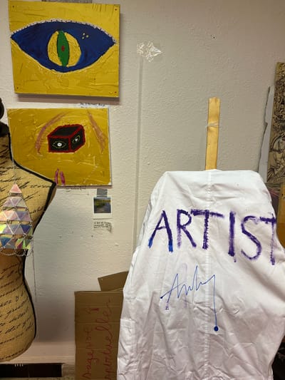 The image shows a corner of what appears to be an artist's studio. On the left, a mannequin is draped in a shimmering, geometric top made of iridescent triangular elements. Behind the mannequin, two square paintings hang on a textured white wall. The top painting is yellow with a large, stylized blue and green eye. The bottom painting is also yellow and features a black cube with eyes. Below these paintings are several small pieces of paper, including a polaroid photograph of a landscape.

To the right, a white garment, possibly a smock or a sheet, is draped over an easel. The word "ARTIST" is written in large, bold purple letters across the garment, and below it is a signature in blue ink. A light-colored wooden stick, likely part of the easel, protrudes from the top of the garment. Further to the right, partially visible, is a piece of artwork with intricate black and white illustrations, featuring elements of mythology or fantasy. At the bottom right, a cardboard box is visible, with handwritten text in pink.

The overall environment is cluttered, suggesting a working creative space. There is no information in the image to indicate the location is Marseille, France.