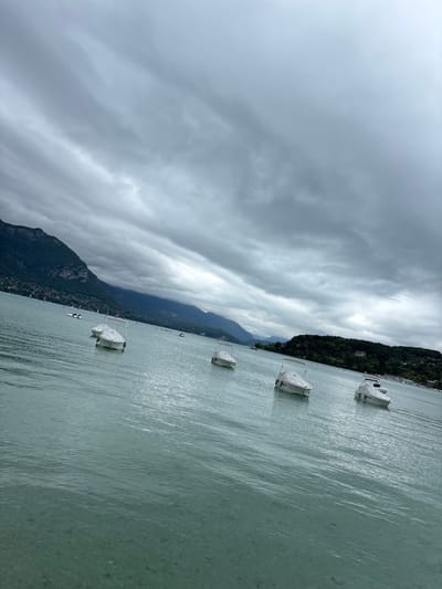 The image depicts a serene scene on Lake Annecy, in Annecy, France, under a cloudy sky. Several white motorboats are moored or gently drifting on the tranquil, turquoise waters. In the background, the lush, green slopes of the French Alps rise majestically, with a hint of the town of Annecy visible along the shoreline, characterized by clusters of buildings and trees. The water is clear enough to reveal the rocky bottom in the foreground, creating a sense of depth and pristine nature. The overall atmosphere is peaceful and contemplative, with the muted colors of the landscape and sky contributing to a subdued, yet beautiful, ambiance.
