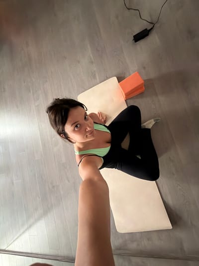 A woman in a light green sports bra and black leggings is sitting on a yoga mat on a light gray laminate floor. She is holding a phone in her right hand, taking a selfie. Two orange yoga blocks are stacked on top of the yoga mat to her right. A black power adapter with a cord is on the floor to the left of the yoga blocks. The scene appears to be indoors, likely in a residential setting within Vracar Municipality, Serbia.